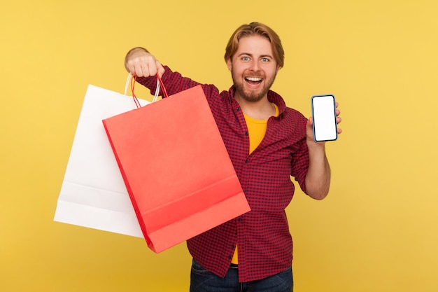 Happy handsome shopper guy in checkered shirt holding shopping bags and mobile phone blank mock up display, looking at camera with excitement toothy smile. studio shot isolated on yellow background