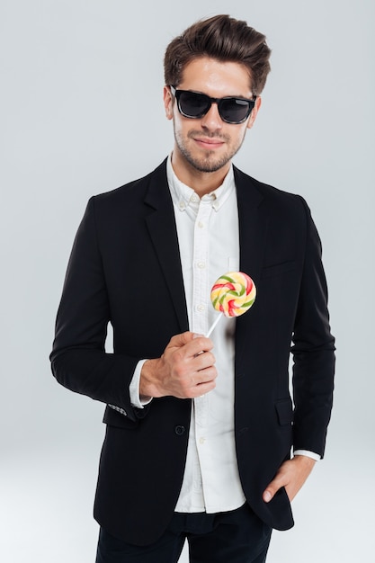 Happy handsome stylish man in sunglasses and black suit holding colorful lollipop over grey wall