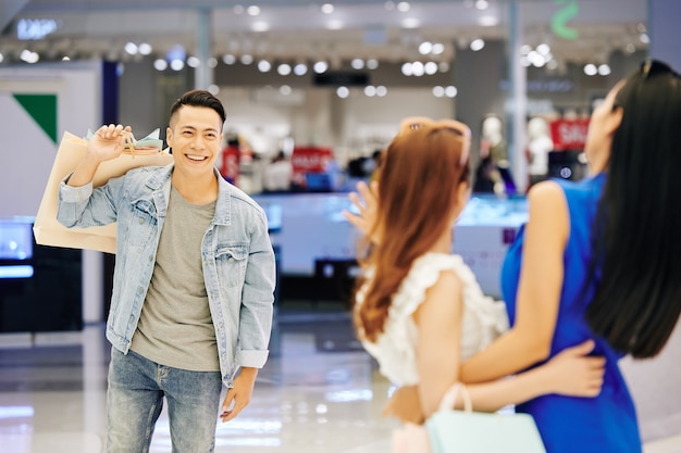 Happy handsome young Asian man with shopping bag walking to his female friends waiting for him in shopping mall