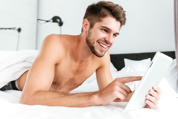 Happy handsome young man smiling and using tablet in bed