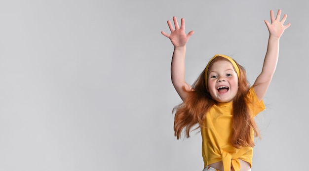 Happy happy little redhaired girl in orange tshirt rejoices\
with raised hands showing happiness and satisfaction emotions\
cheerfully standing against studio wall background banner