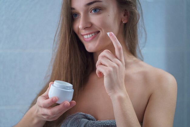 Happy healthy attractive brunette young woman in towel apply moisturizing cream to face in bathroom after shower at home