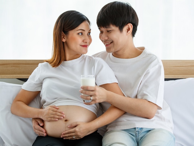 행복하고 건강한 가족 개념입니다. 함께 침대에 앉아 있는 젊은 부부의 그림. 젊은 남편이 웃고 우유 한 잔을 들고 사랑으로 임신한 아내에게 줍니다.