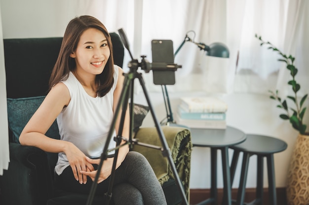 Happy healthy smiling young Asian woman blogger looking at camera while using smartphone for recording live video vlog at home