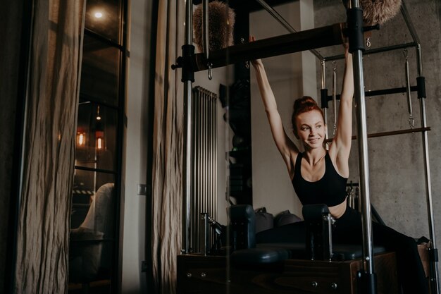 Happy healthy woman in black sportswear stretching upper body pulling up by hands on frame of cadillac reformer during pilates training in gym or studio, looking aside and smiling. Active lifestyle