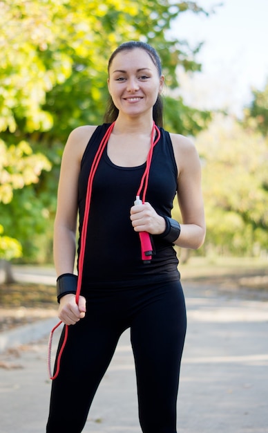 Happy healthy woman with a skipping rope around her neck and a fit shapely body standing on a road in a park