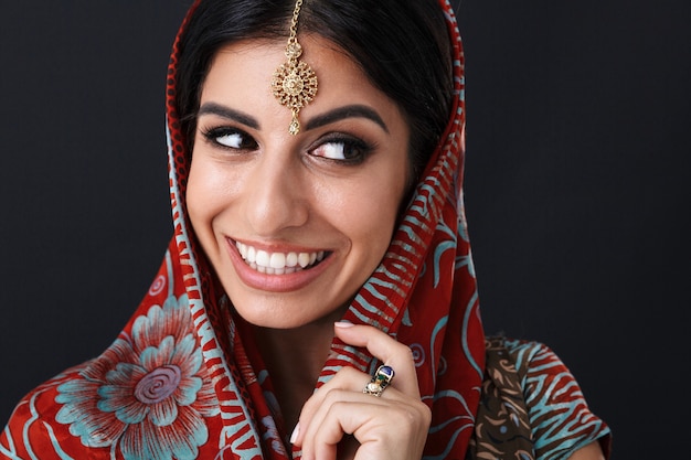 happy hindus girl wearing traditional indian saree dress and ethnic jewelry covering her head with dupatta scarf isolated over black wall