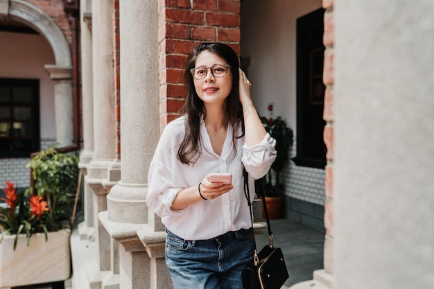 happy hipster asian japanese girl dressed in urban style leans against brick pillar with smart phone in hands. young elegant woman in glasses smiling holding mobile phone and search online guide book