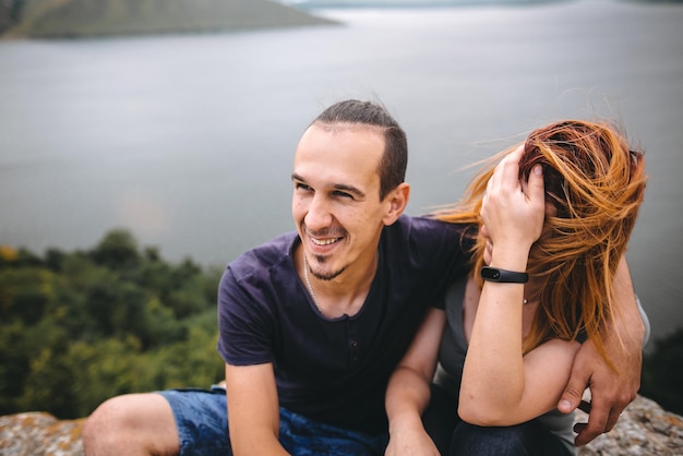 Happy hipster couple sitting and laughing on top of rock mountain with beautiful view on river Tourist couple hugging and smiling on windy cliff Traveling together Copy space