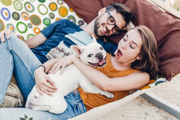 Photo happy hipster couple with bulldog relaxing in hammock on the beach in sunset light summer vacation stylish family with funny dog resting cute moments space for text