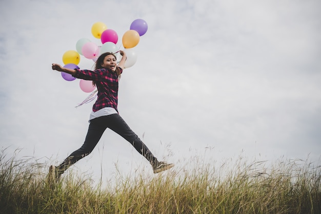 Foto felice ragazza hipster che salta con palloncini giocattolo colorato all'aperto. giovane donna che ha divertimento nel campo verde contro il cielo blu. concetto di lifestyle di libertà delle donne.