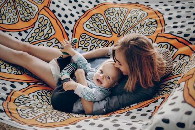 Happy hipster mother playing with her cute little son on hammock in summer sunny park stylish mom with kid relaxing in forest and cuddling having fun space for text