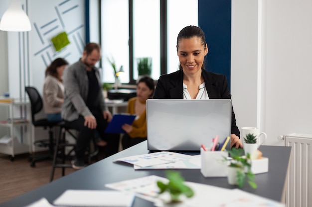 Happy hispanic lady typing on laptop sitting at desk in start up business office drinking coffee while diverse colleagues working in background