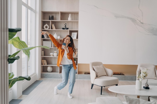 Photo happy hispanic woman singing at home with mobile phone as with microphone online music listening