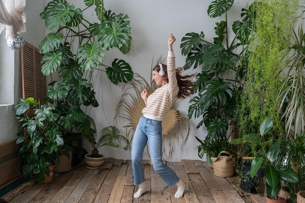 Happy hispanic woman wear wireless headphones relaxing dancing in home garden with tropical plants