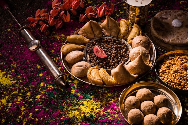 Поздравительная открытка Happy Holi, показывающая индийские традиционные сладкие и соленые блюда, цветы и порошковые цвета, расположенные на белом фоне или глине. Выборочный фокус