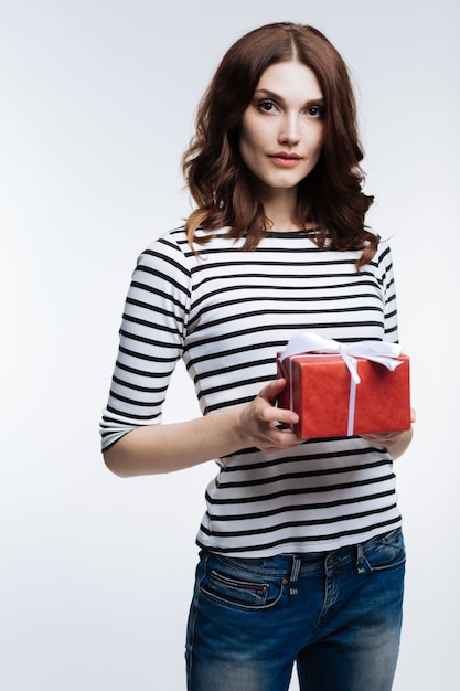 Happy holiday. Charming auburn-haired young woman in a striped pullover holding a red gift box tied up with a bow while posing on grey background