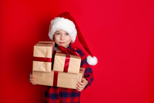 幸せな休日とメリークリスマス。赤い背景に彼の手に贈り物を持った帽子をかぶった少年の肖像画。テキストの場所。高品質の写真