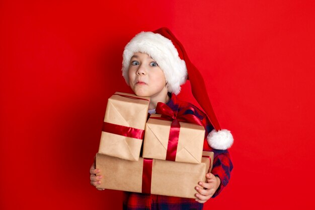 Happy holiday and Merry Christmas. Portrait of a boy in a cap with gifts in his hands on a red background. A place for text. High quality photo