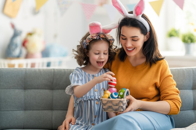 Happy holiday! Mother and her daughter with painting eggs. Family celebrating Easter. Cute little child girl is wearing bunny ears.