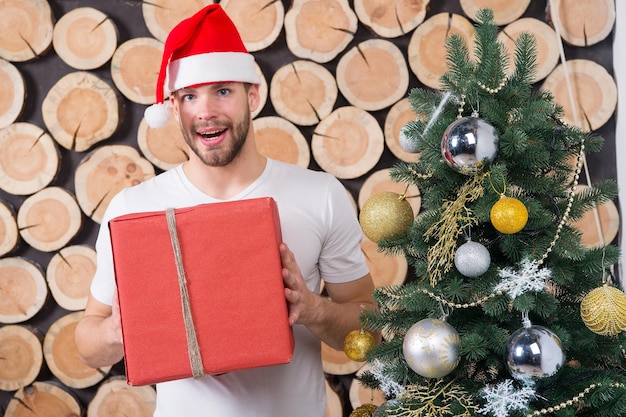 幸せな休日のお祝い。クリスマスツリーのボックスを持つサンタの男。クリスマスと新年は準備を提示します。赤い帽子のマッチョは贈り物と笑顔。ボクシングデーのコンセプト。