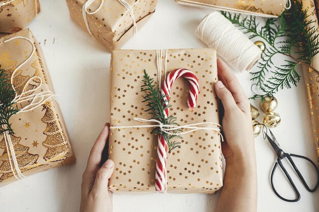 写真 スタイリッシュなクリスマス ギフトをラッピング幸せな休日手フラット レイアウト白い木にキャンディー杖お祝い紙黄金の装飾組成物でモダンなクリスマス ギフト ボックスを準備する人