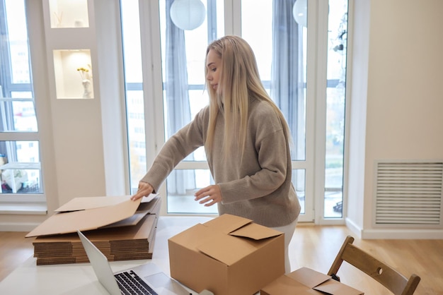 Happy homeowner renter girl using laptop at pile of stacked boxes ordering delivery shipping transpo