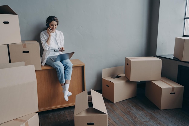 Happy homeowner talking on phone Hispanic girl is sitting among carton boxes and working on pc