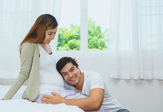 Happy husband listening to baby in belly of pregnant wife in bedroom at home