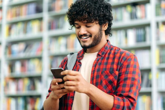 Happy indian or arabian millennial guy freelancer or student\
using his smartphone chatting with friends on social networks\
texting sms browses internetsmile friendly wireless technology\
concept