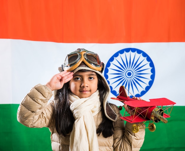 Happy Indian or asian girl kid flying a toy metal airplane or saluting against indian flag background - Kid and flying for country