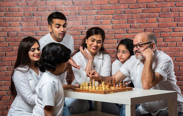 Happy Indian asian multigenerational family of six playing chess or Shatranj which is a popular board game, wearing white cloths against red brick wall