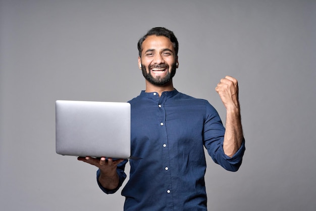 Happy indian business man holding laptop isolated on gray celebrating result
