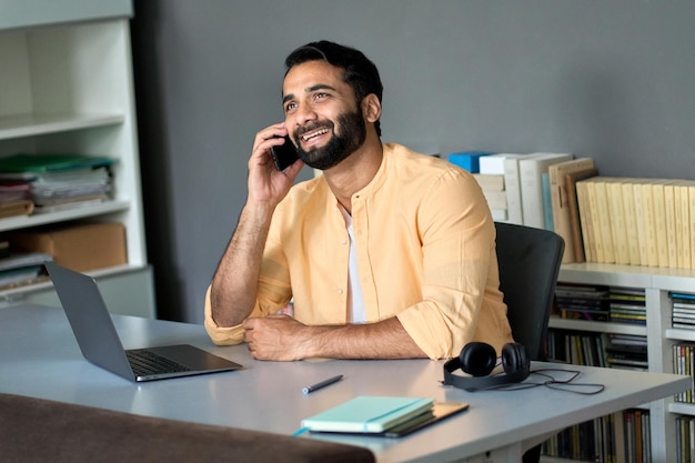 Happy indian business man sitting at work desk talking on phone\
making call