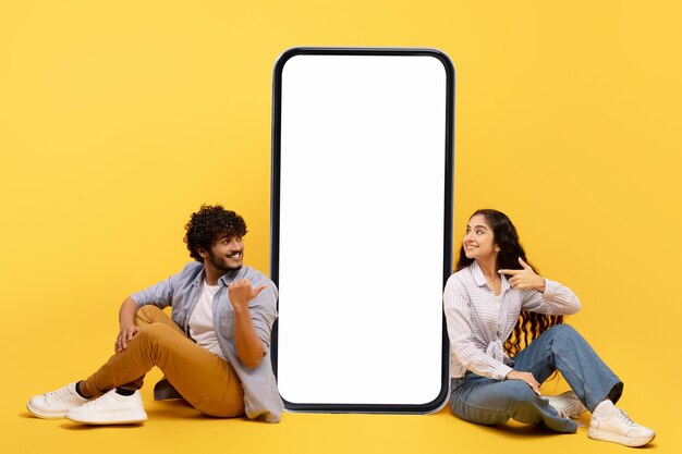 Happy indian couple sitting near big smartphone with blank white screen demonstrating copy space for