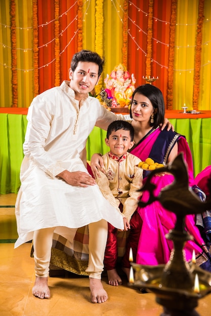 Happy Indian Family Celebrating Ganesh Festival or Chaturthi - Welcoming or performing Pooja and eating sweets in traditional wear at home decorated with Marigold Flowers