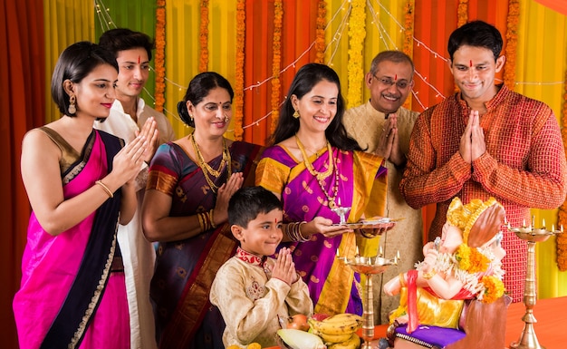 Happy Indian Family Celebrating Ganesh Festival or Chaturthi - Welcoming or performing Pooja and eating sweets in traditional wear at home decorated with Marigold Flowers