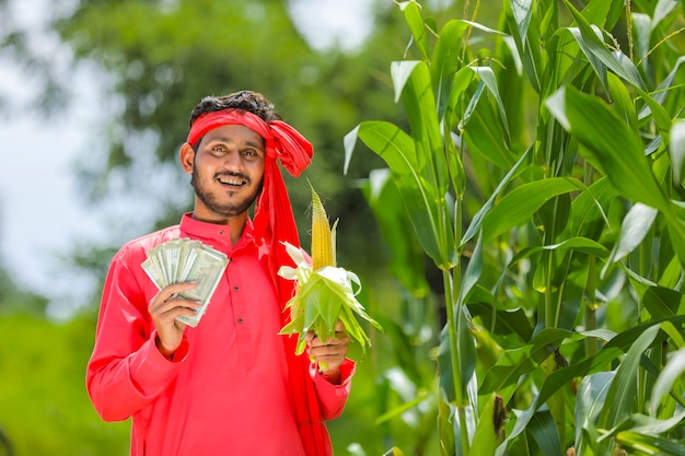 Coltivatore indiano felice che mostra frutta di mais e valuta indiana al campo di mais verde