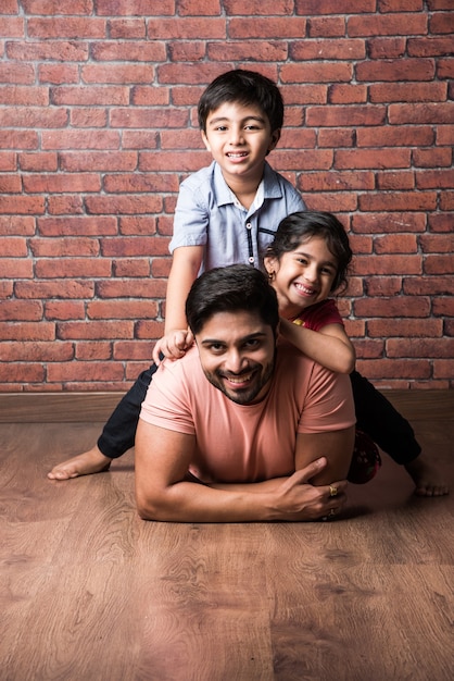 Happy Indian girl and boy playing riding on back or piggyback with father, indoor shot with mother looking