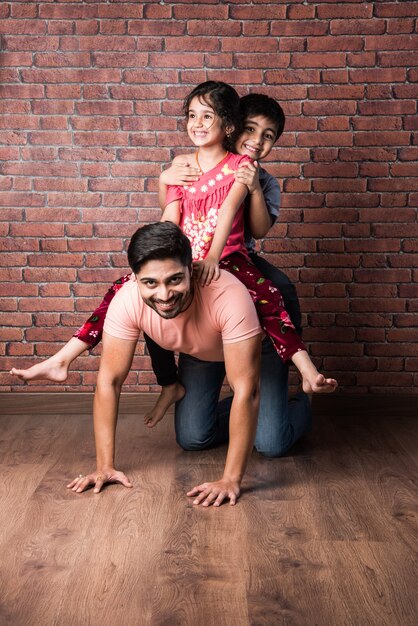 Happy Indian girl and boy playing riding on back or piggyback with father, indoor shot with mother looking