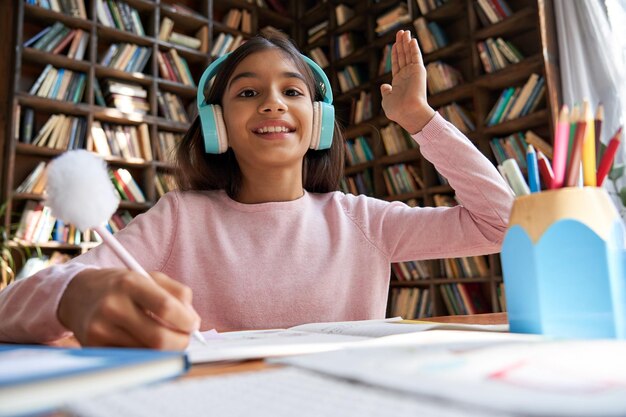 写真 ヘッドフォンを着けた幸せなインドのラテンプレティーンの女の子の学校の生徒が、自宅のウェブカメラビューでビデオ会議通話による教師の家庭教師との仮想レッスンクラスでオンライン遠隔学習で手を上げる