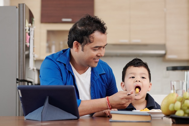 행복한 인도 남성이 디지털 태블릿으로 식탁에 앉아 있을 때 아들에게 망고와 바나나 조각을 먹입니다
