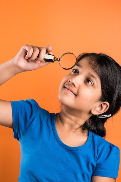 Happy indian school girl child with magnifying glass, standing isolated over orange background