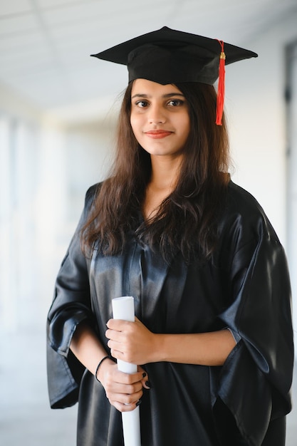 Happy Indian university student in graduation gown and cap holding diploma certificate.