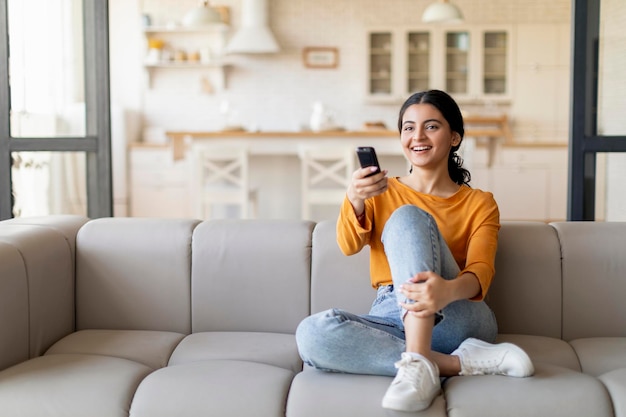 Happy indian woman with remote controller in hand watching tv at home