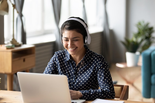 Happy indian young girl student wear headphone watch webinar\
listen online course communicate by conference video call e learn\
language in app laugh study with teacher lesson look at laptop at\
home