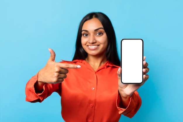 Happy indian young lady showing cellphone empty screen blue background