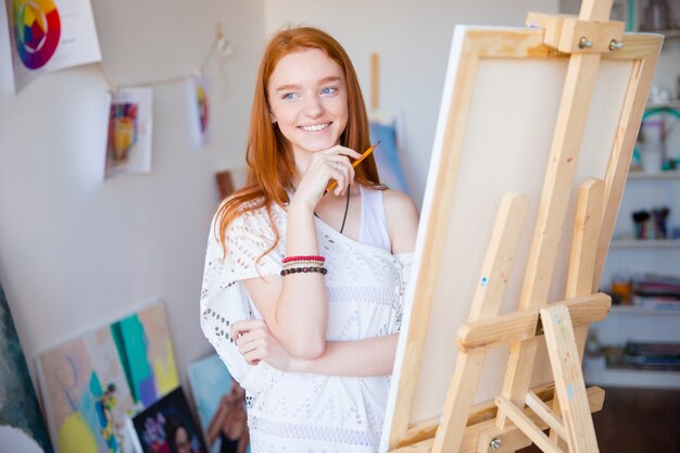 Happy inspired smiling female artist with long red hair drawing with pencil in