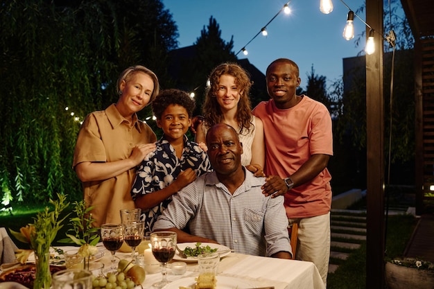 Happy intercultural family of five young and mature people in casual attire looking at camera while senior man sitting against four other members