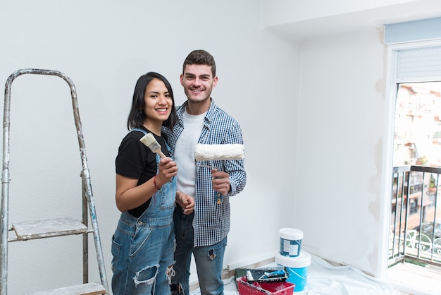 Happy interracial young couple looking at camera holding painting tools to renovating the living room at his new apartment.